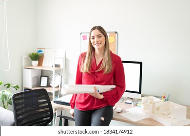 Good Looking Female Architect Holding A Few Rolled Up Plans While Sitting On A Desk In An Office And Smiling