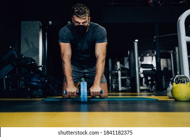 Good Looking Handsome Male Athlete With Protective Face Mask Exercising In Modern Fitness Gym. Dark Muddy Light With Strong Shadows. Pandemic Sports Indoors Concept.