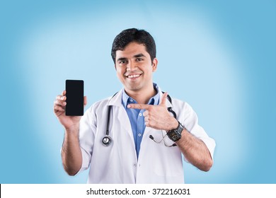 Good Looking Indian Male Doctor Using/speaking Over Or Presenting Smartphone, Standing Isolated Over Light Blue Colour.