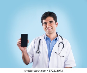 Good Looking Indian Male Doctor Using/speaking Over Or Presenting Smartphone, Standing Isolated Over Light Blue Colour.