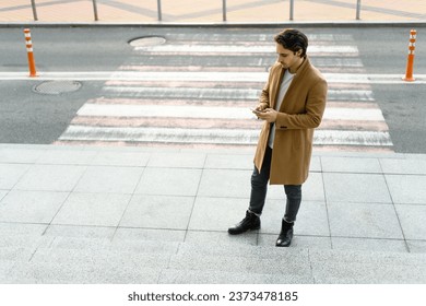 Good looking man in brown coat using smartphone near crosswalk on urban street	
