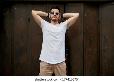 Good Looking Man Posing In Front Of A Rusty Dark Metal Door Wearing Sunglasses, White T-shirt And Khaki Cargo Pants Holding Hands Behind His Head. 