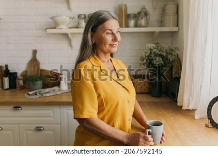 Good looking mature woman enjoying cup of tea on kitchen. Senior mother waiting for child, looking out of window. Parents house, sweet home, family, waiting, mothers love concept