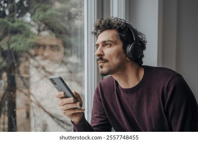 Good looking middle eastern man with on-ear headphones listening to music and relaxing near a window.