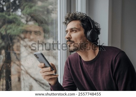 Good looking middle eastern man with on-ear headphones listening to music and relaxing near a window.