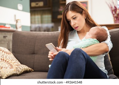 Good looking mother taking care of her newborn baby while using her smartphone to read some emails and texts