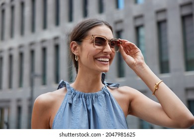 Good looking positive woman enjoys sunny day wears sunglasses and blue dress smiles positively poses against blurred background in ancient city. Happy young female model strolls in urban place - Powered by Shutterstock