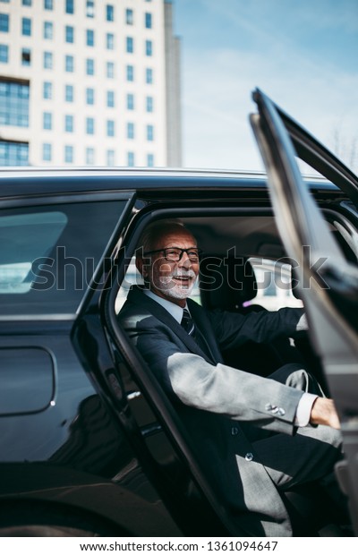 Good looking senior business man sitting on\
backseat in luxury car. He opens car doors and going or stepping\
out. Big busness bulding in background. Transportation in corporate\
business concept.