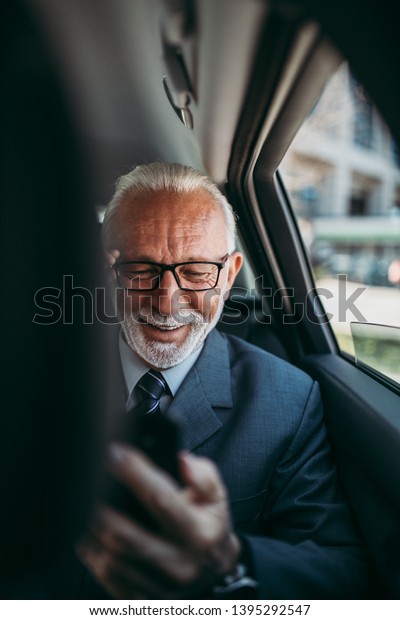 Good looking senior business\
man sitting on backseat in luxury car. He using his smart phone and\
reacting emotionally. Transportation in corporate business\
concept.