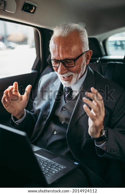 Good looking senior business\
man sitting on backseat in luxury car. He using his laptop and\
reacting emotionally. Transportation in corporate business\
concept.