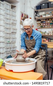 Good Looking Senior Female Potter Working On Pottery Wheel While Sitting  In Her Workshop