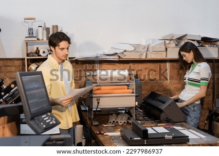 good looking typographer looking at printed paper near pretty colleague