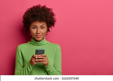 Good looking woman with Afro hair types message on smartphone, browses network, looks confidently at camera, has online communication via special application, wears green poloneck. Mobile services - Shutterstock ID 1626507640