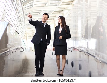 Good Looking Young Asian Business Man And Woman Wearing Black Suits, Walking  In Public Walkway , Talking And Pointing Outside.