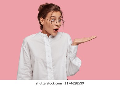 Good Looking Young Caucasian Woman In Round Transparent Eyewear, Keeps Hand Raised, Dressed In Casual Outfit, Pretends Holding Something Wonderful, Isolated Over Pink Background. Look There!