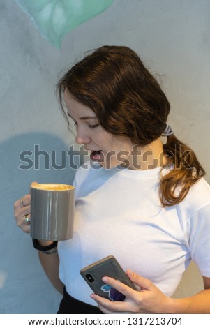 Good looking young hipster girl in stylish clothes and shades messages online and checks newsfeed on smart phone, sits at coffee shop alone, looks out of window, enjoys free wifi during lunch