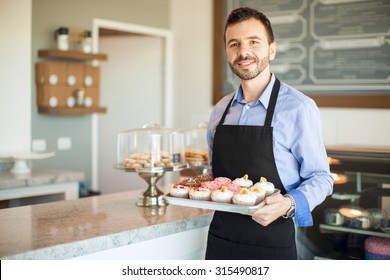 Good Looking Young Hispanic Man Holding A Tray Of Cupcakes And Greeting Customers To His Cake Shop