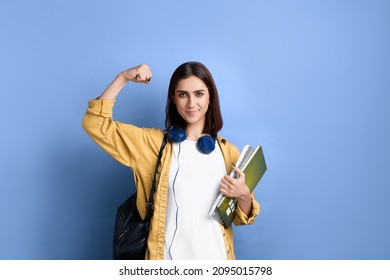 Good luck. Strong smiling student girl shows muscles, passed a milestone, confident in her knowledge, holding books, wearing yellow shirt, white t-shirt, black bag and headphones over neck