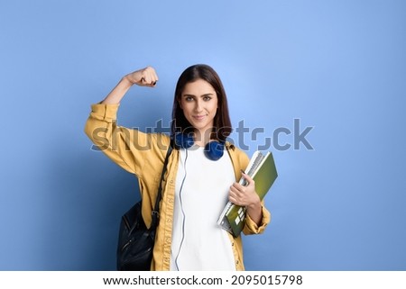 Good luck. Strong smiling student girl shows muscles, passed a milestone, confident in her knowledge, holding books, wearing yellow shirt, white t-shirt, black bag and headphones over neck