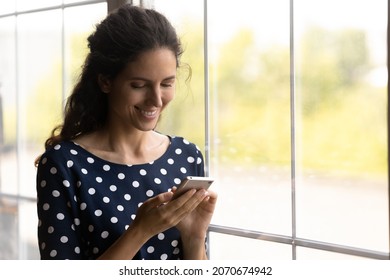 Good Message. Happy Young Latina Female Read Positive News On Cell Phone Screen Check Social Network Account Buy Goods Online. Smiling Woman Stand By Big Light Window At Home Use Telephone. Copy Space