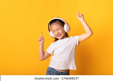 Good Mood. Adorable Asian Kid Listening To Music Wearing Wireless Headset And Dancing, Isolated Over Yellow Wall