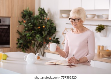 In A Good Mood. Cheerful Nice Elderly Woman Making Notes And Drinking Tea While Smiling In The Kitchen