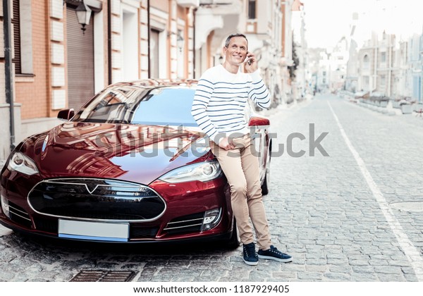 Good mood. Positive\
delighted male keeping smile on his face and leaning on his auto\
while having dreamy mood