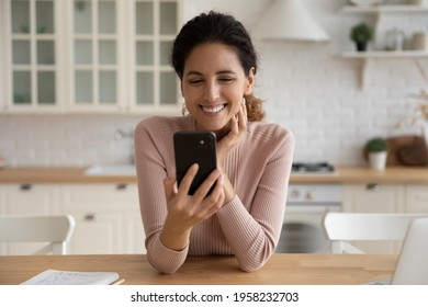 In a good mood. Smiling young hispanic lady sit at kitchen table enjoy talk by video call dating on website read positive news in email. Cheerful latina woman spend time at social network in phone app