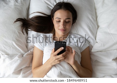 Good morning from afar! Calm smiling young female lying in comfy bed at home or in hotel suite after waking up spending time with mobile phone texting messaging making answering call reading good news