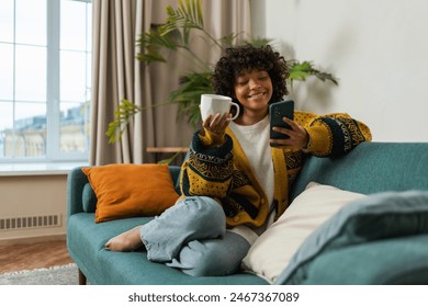 Good morning. African girl drinking coffee holding smartphone sitting on couch at home Woman with cell phone surfing internet using social media apps. Shopping online Internet news cellphone addiction - Powered by Shutterstock