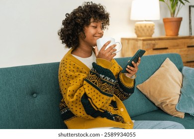 Good morning. African girl drinking coffee holding smartphone sitting on couch at home Woman with cell phone surfing internet using social media apps. Shopping online Internet news cellphone addiction