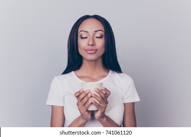 Good morning! Close up portrait of charming dreamy african young woman, drinking hot tea. She is relaxed, in casual white wear, with closed eyes