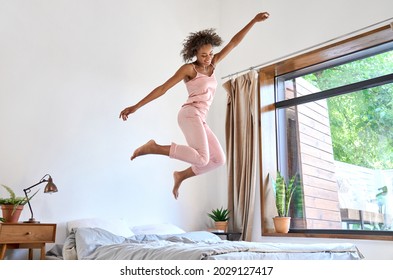 Good morning concept. Happy carefree positive playful young African American woman wearing pajamas jumping dancing on bed waking up flying in air having fun feeling joy alone in cozy bedroom. - Shutterstock ID 2029127417