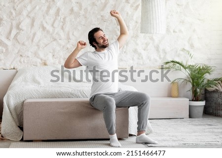 Good morning. Happy bearded young man waking up in bed and stretching his arms, smiling with closed eyes, wearing pajamas and socks, panorama with copy space, bedroom interior