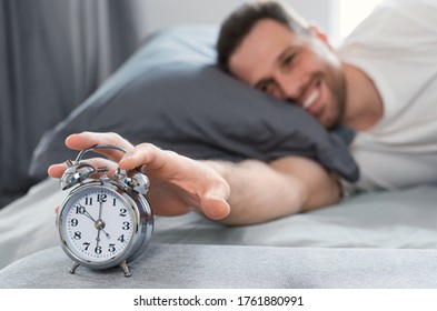 Good Morning. Happy Well-Slept Man Turning Off Alarm-Clock Waking Getting Up Lying In Bed At Home. Shallow Depth, Selective Focus