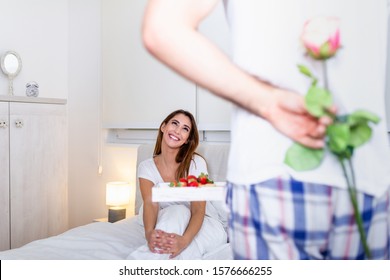 Good morning! Healthy breakfast in bed. Beautiful young woman is lying in bed while her handsome man bringing tasty breakfast and rose behind his back.
