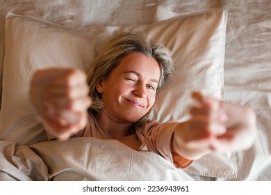 Good morning, new day, weekend, holiday. Happy middle aged woman sits on bed, lady stretching arms after sleep and enjoying morning in cozy comfort bedroom interior, free space