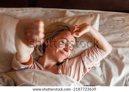 Good morning, new day, weekend, holiday. Happy middle aged woman lying on bed, lady stretching arms after sleep and enjoying morning in cozy comfort bedroom interior, free space