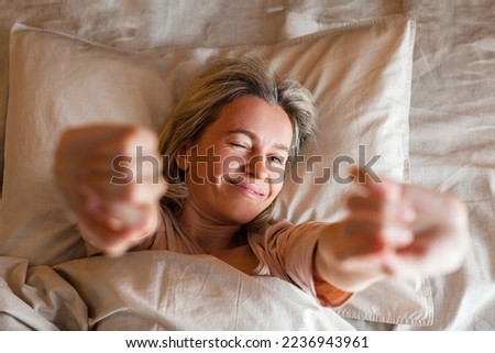 Good morning, new day, weekend, holiday. Happy middle aged woman sits on bed, lady stretching arms after sleep and enjoying morning in cozy comfort bedroom interior, free space