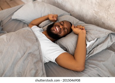 Good Morning. Satisfied young African American man stretching arms after wake up, feeling rested, happy bearded black guy sleeping lying on the gray linen flax bedsheets at home, relaxing in bedroom