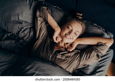 Good morning. View from above of young beautiful relaxed brunette female with closed eyes in satin pajamas waking up in her bed fully rested, happy woman stretching after night sleep