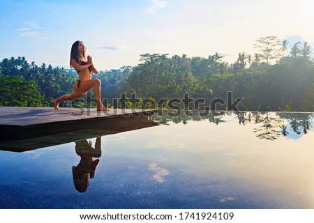 Good morning with yoga meditating on sunrise background. Active woman in bikini practicing at villa poolside to keep fit and health. Woman fitness training, sport activity on summer family holiday.