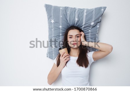 Good morning. Young woman drinking coffee with a pillow under her head, light gray background