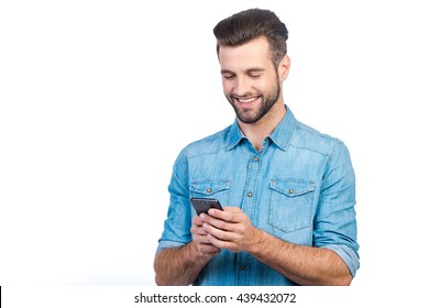 Good News From Friend. Confident Young Handsome Man In Jeans Shirt Holding Smart Phone And Looking At It With Smile While Standing Against White Background 