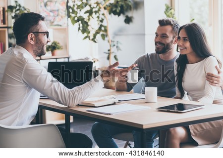 Good news! Happy young couple bonding to each other and listening to man sitting at the desk in front of them and gesturing