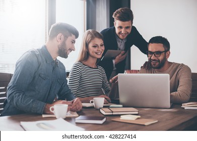 Good News For Us. Young Handsome Man Looking At Laptop With Smile While Sitting At The Office Table On Business Meeting With His Coworkers