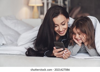 Good Night Or Good Morning. Young Beautiful Mother And Her Little Daughter Use The Phone While Lying On The Bed At Home And Smiling At The Camera.