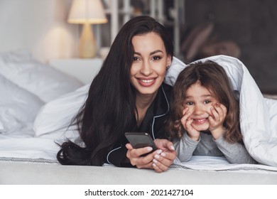 Good Night Or Good Morning. Young Beautiful Mother And Her Little Daughter Use The Phone While Lying On The Bed At Home And Smiling At The Camera.