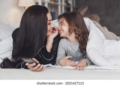 Good Night Or Good Morning. Young Beautiful Mother And Her Little Daughter Use The Phone While Lying On The Bed At Home And Smiling At The Camera.