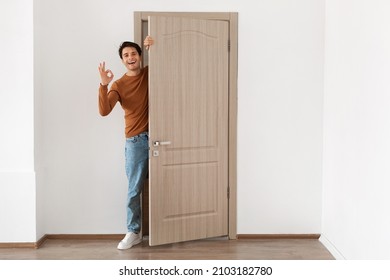 Good Offer. Cheerful young guy standing in doorway of his apartment, smiling millennial male homeowner holding ajar door looking peeping out greeting visitor, showing ok sign gesture, full body length - Powered by Shutterstock